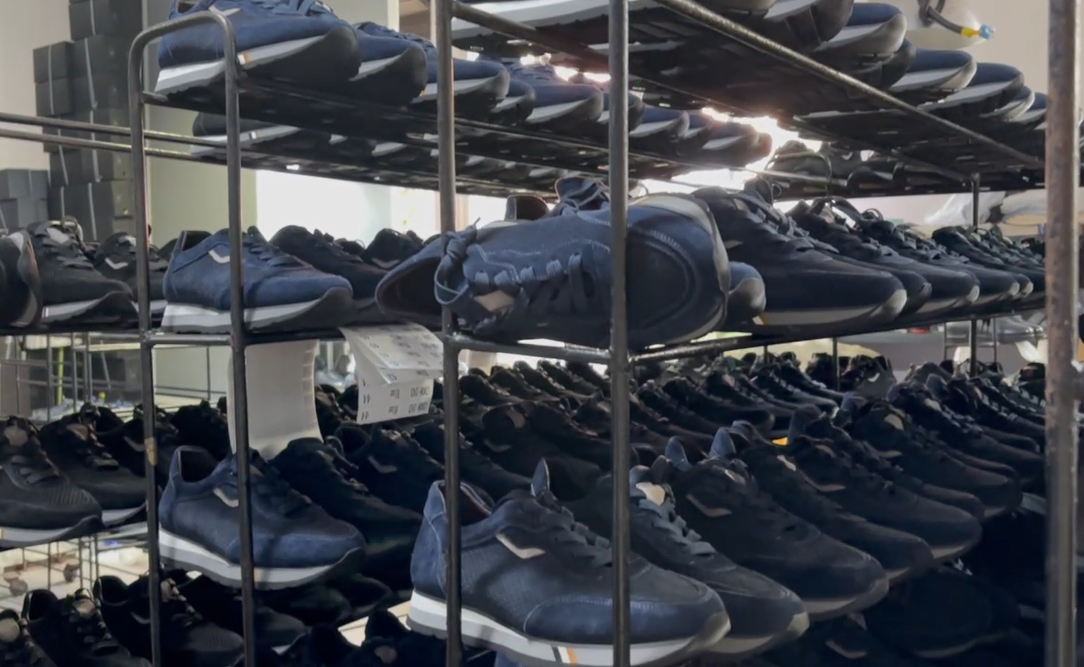 Bulk dark blue and black athletic sneakers stacked on multi-tier metal storage racks in a shoe factory warehouse, demonstrating large-scale production capacity and ready-to-ship wholesale inventory.