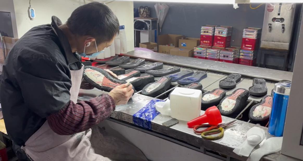 Close-up of a masked factory worker manually applying adhesive to black rubber shoe soles on a workshop production line, demonstrating precise craftsmanship and quality control in a wholesale footwear factory.