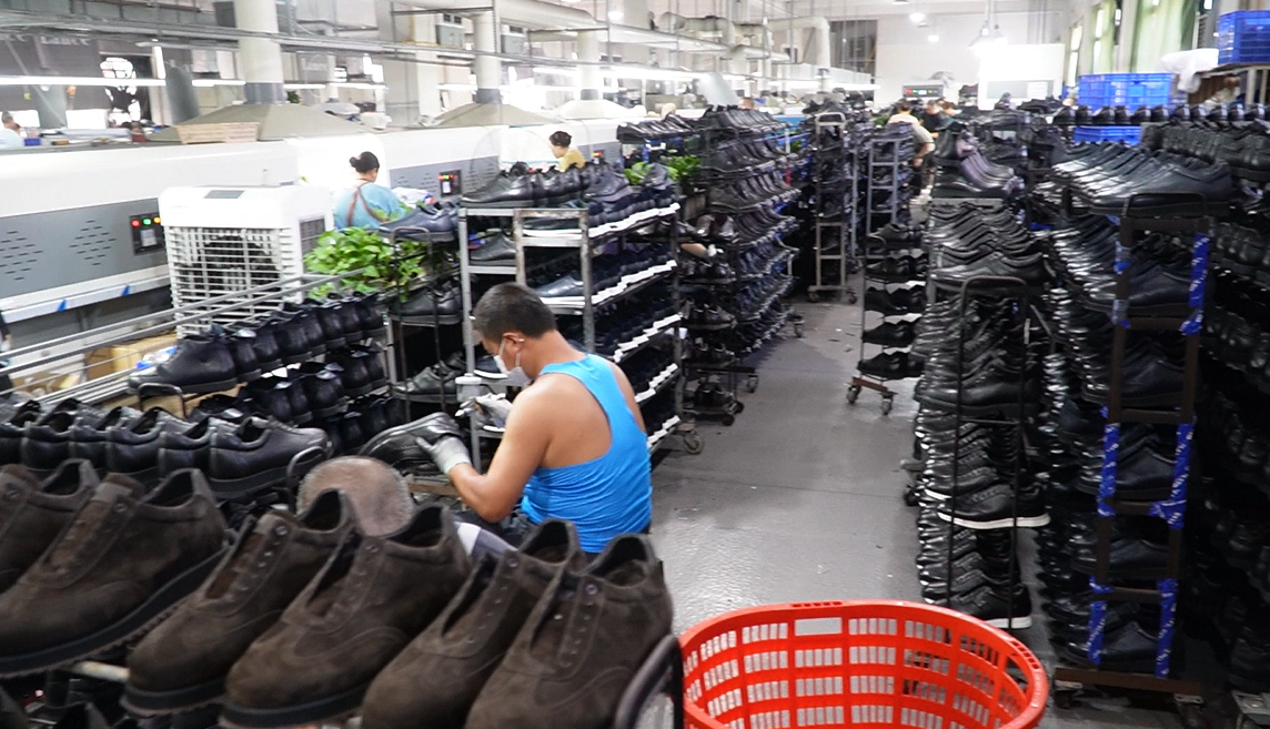 Wide shot of a busy shoe manufacturing workshop with massive quantities of black leather shoes stacked on rolling racks, and a worker polishing a shoe at a workstation, demonstrating strong mass production capacity.