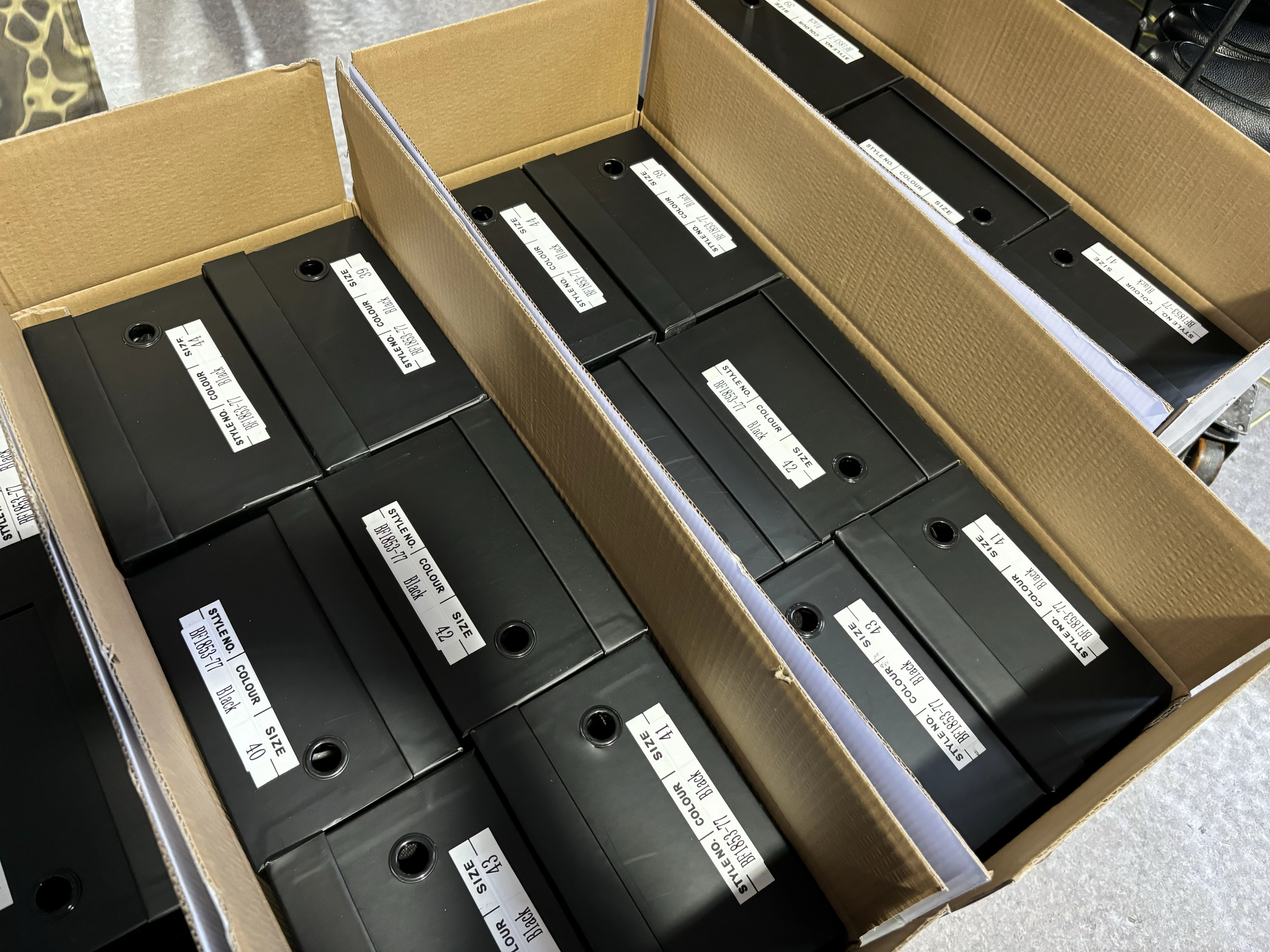 Packaged black shoe boxes with style, color, and size labels neatly organized inside large corrugated cardboard cartons, demonstrating efficient wholesale packaging, inventory management, and export preparation at the shoe factory